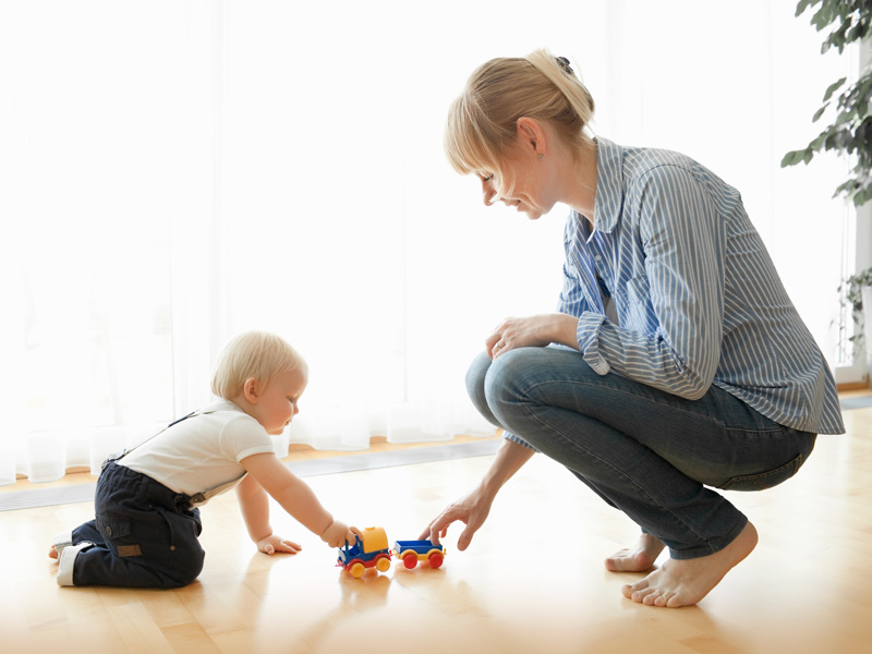 Eine blonde Mutter spielt mit ihrem kleinen Kind gemeinsam auf einem Holzboden mit einem bunten Spielzeugauto