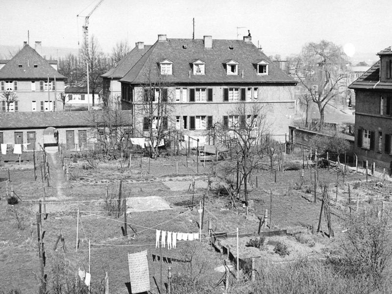 GSG_90Jahre_Engerserland_Innenhof_800x600px_sRGB Schwarzweißfoto eines Innenhofs mit alten Wohngebäuden und Wäscheleinen mit hängender Kleidung