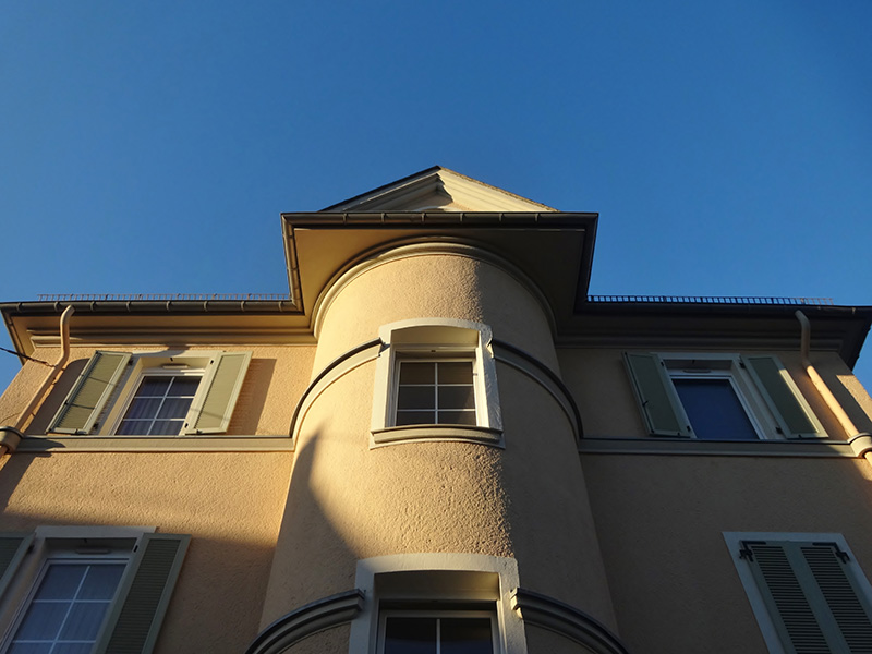 Fassade eines beigen Hauses mit rundem Erker und Fensterlaeden vor einem klaren blauen Himmel