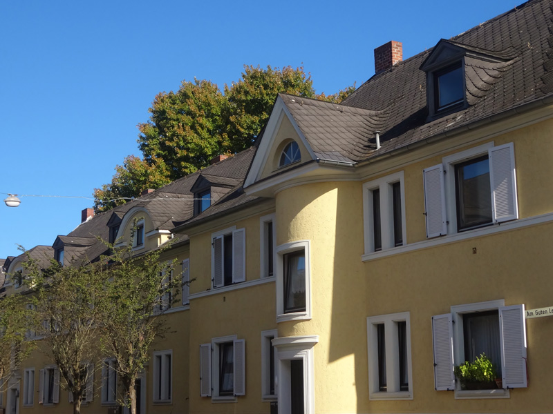 Gelbes Wohnhaus mit Gauben und weißen Fensterläden unter blauem Himmel vor grünen Bäumen