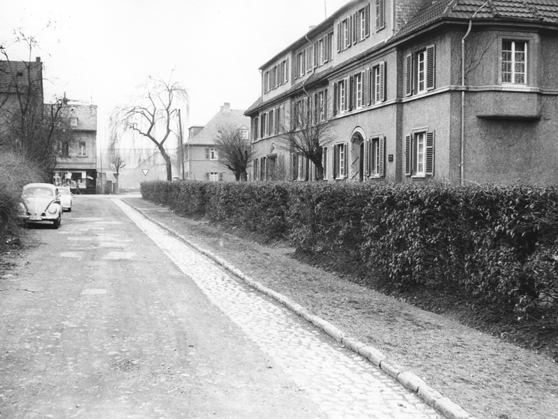 GSG_90Jahre_Sonnenstrasse_800x600px_sRGB Schwarzweißfoto einer ruhigen Straße mit alten Wohnhäusern, einer Hecke und einem geparkten VW Käfer