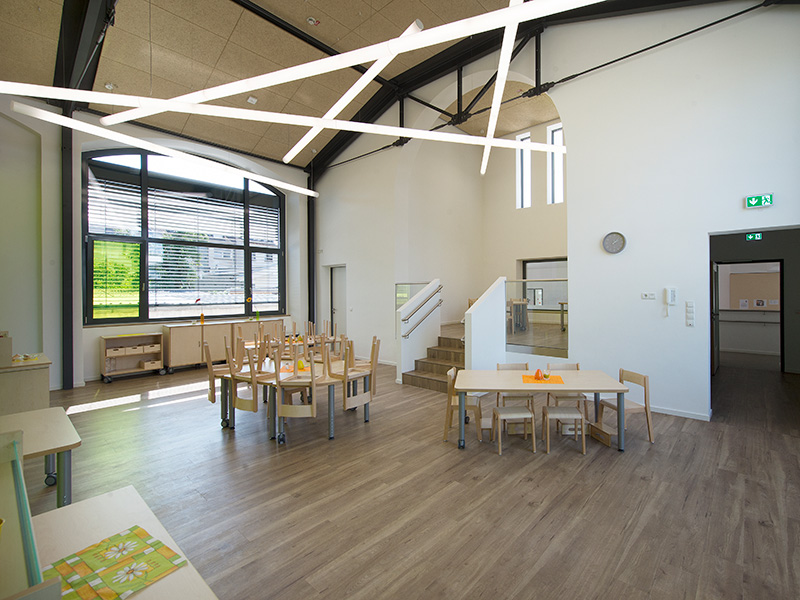 Heller Kindergarten Gruppenraum mit modernen Holztischen, Stühlen und auffälliger moderner Deckenbeleuchtung