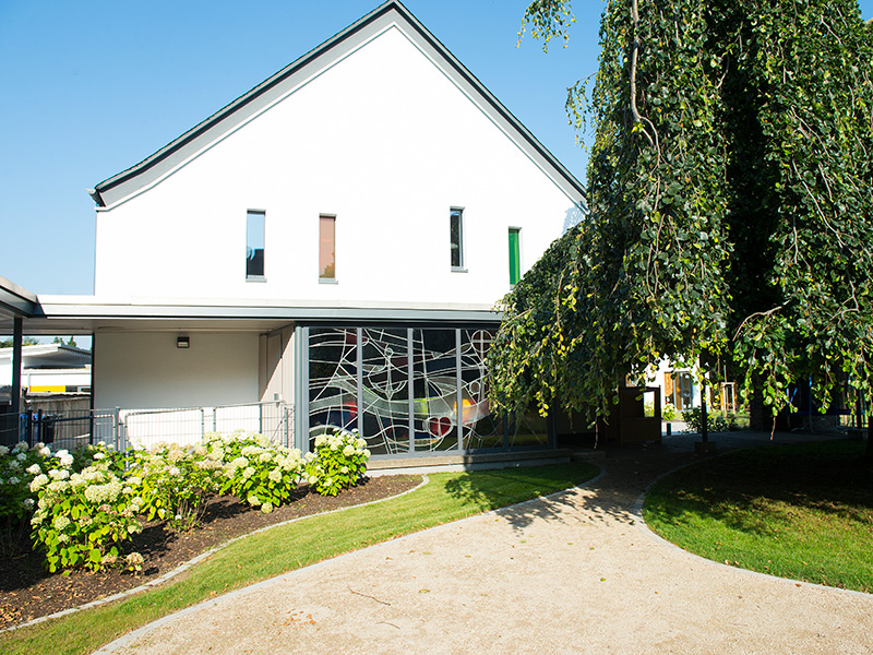 GSG_Kindergarten_04_800x600px_sRGB Modernes Kindergarten Gebaeude mit weisser Fassade, bunten Glasfenstern und einem gepflegten Garten mit grossem Baum