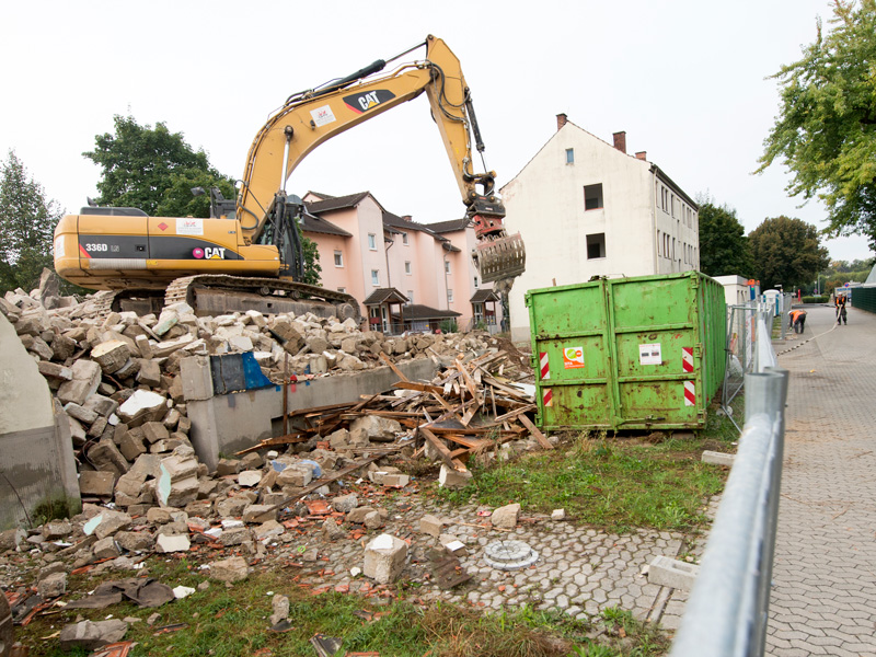 GSG_Stadtvillen_02_800x600px_sRGB Gelber Kettenbagger bei Abbrucharbeiten neben einem grünen Schuttcontainer vor Wohngebäuden