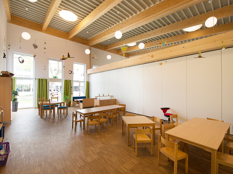 GSG_Kindergarten_06_800x600px_sRGB Heller Kindergarten Gruppenraum mit Holztischen Stühlen und großen Fenstern sowie hängender Dekoration an der Decke