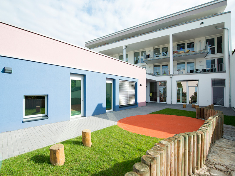 Moderner Kindergarten mit blau rosa Fassade und kleinem Spielplatz mit Rasenfläche und Holzpfosten
