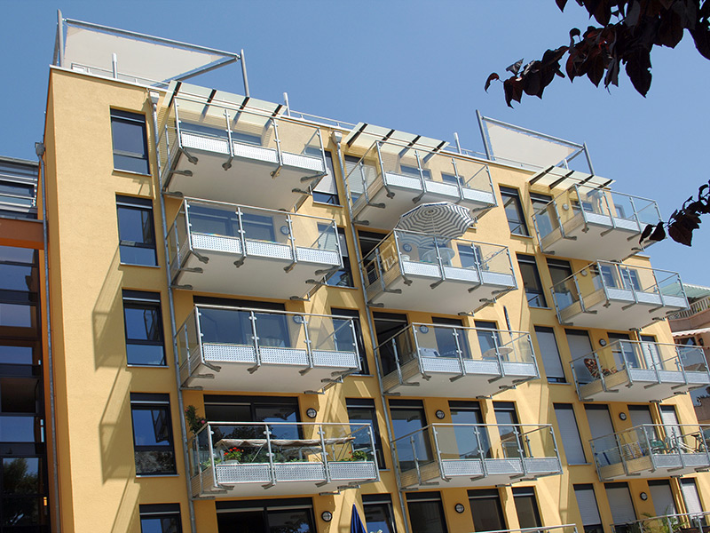 GSG_Gaeste_01_800x600px_sRGB Gelbes Wohnhaus mit Balkonen und Glasgeländern an einer sonnigen Fassade unter blauem Himmel
