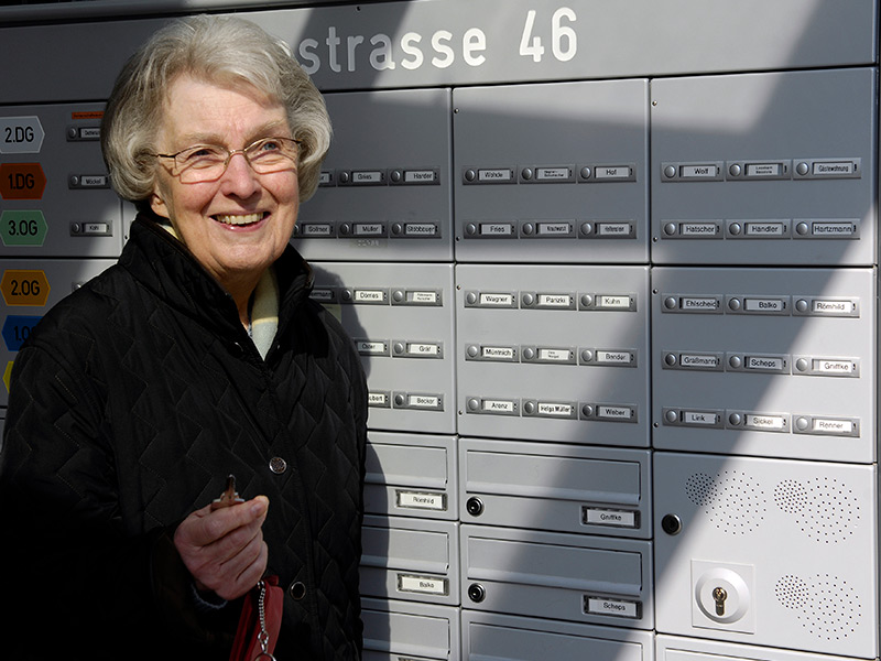 GSG_Senioren_03_800x600px_sRGB Eine lächelnde ältere Dame hält einen Schlüssel vor einer grauen Briefkastenanlage mit Klingelschildern