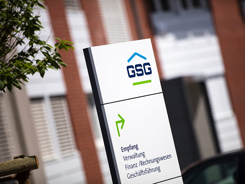 GSG_Wohnungssuche_800x600px_sRGB Weißes Firmenschild der GSG mit Wegweiser zu Empfang Verwaltung Finanzwesen und Geschäftsführung vor einem Gebäude
