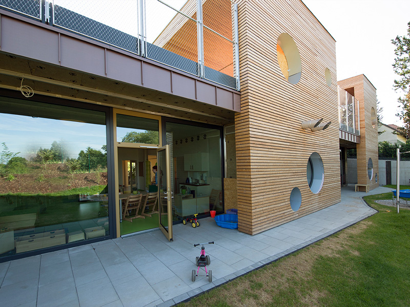 GSG_Kindergarten_08_800x600px_sRGB Moderner Kindergarten mit Holzfassade, runden Fenstern und einer Terrasse mit Spielzeug auf einer Rasenfläche
