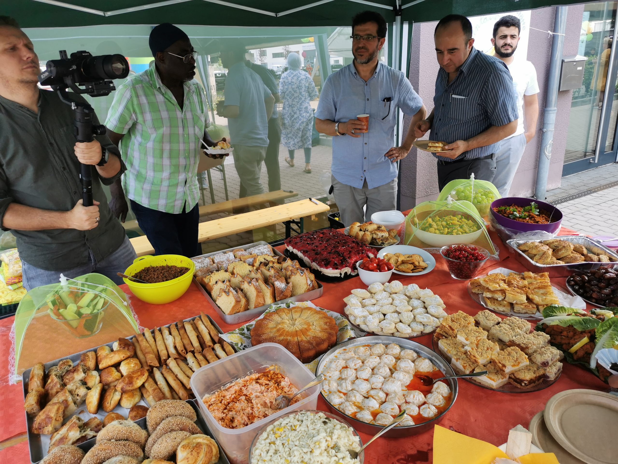 Eine Gruppe von Männern steht an einem reich gedeckten Buffet mit verschiedenen Kuchen, Gebäck und Salaten im Freien