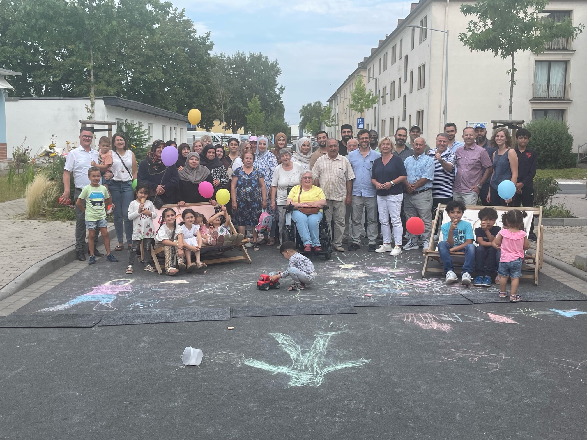 Eine große, diverse Gruppe von Menschen bei einem Straßenfest mit Kreidezeichnungen auf dem Boden und bunten Luftballons