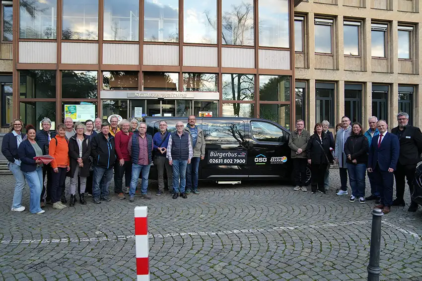 Eine große Gruppe von Menschen steht vor einem schwarzen Bürgerbus mit Firmenlogo vor einem modernen Verwaltungsgebäude