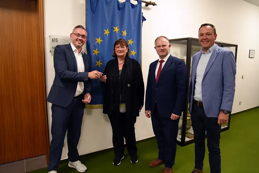 Vier Personen bei einer offiziellen Schlüsselübergabe für einen Bürgerbus vor einer Europafahne