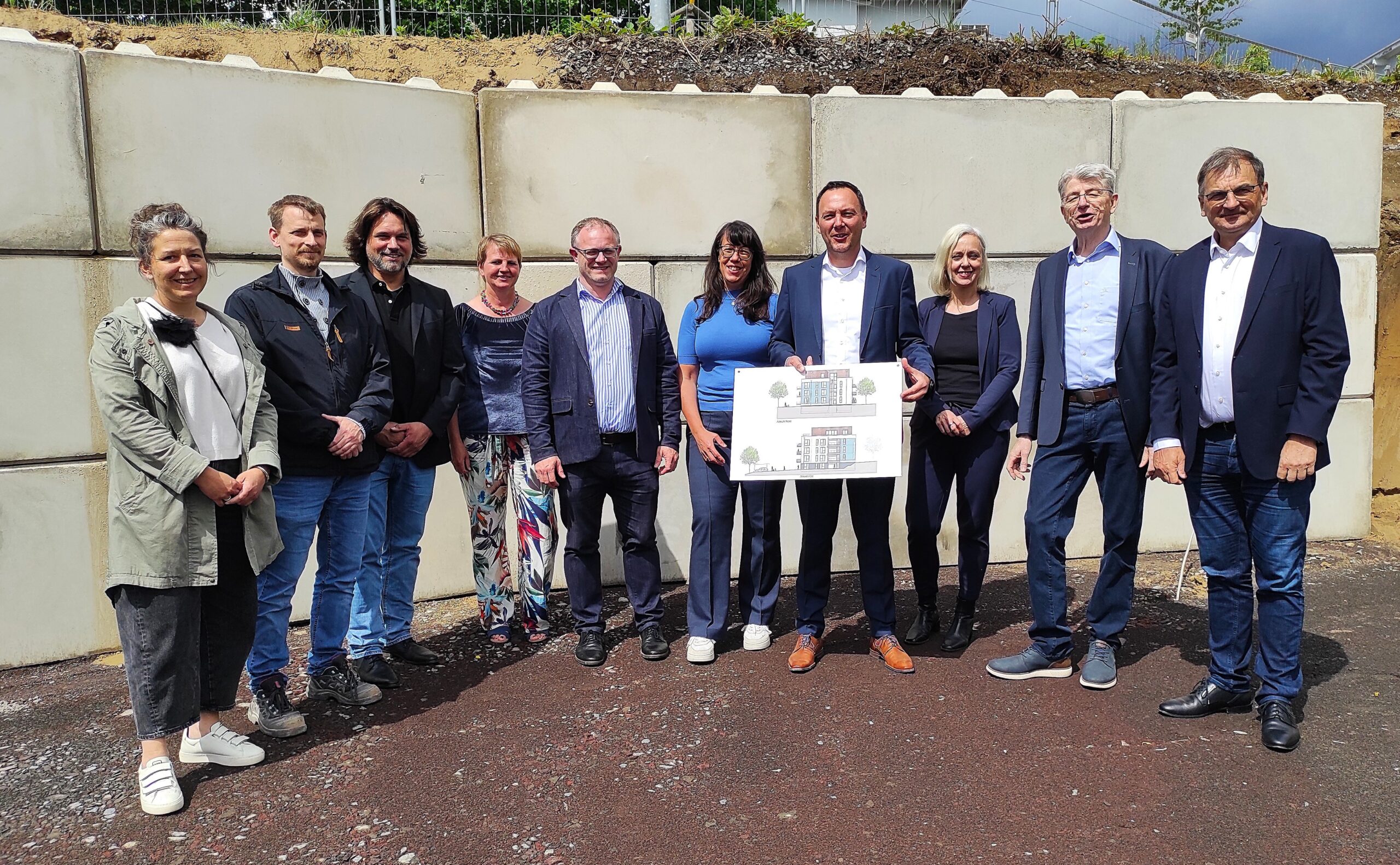 Eine Gruppe von Menschen präsentiert vor einer Betonmauer die Pläne für zwei Neubauten