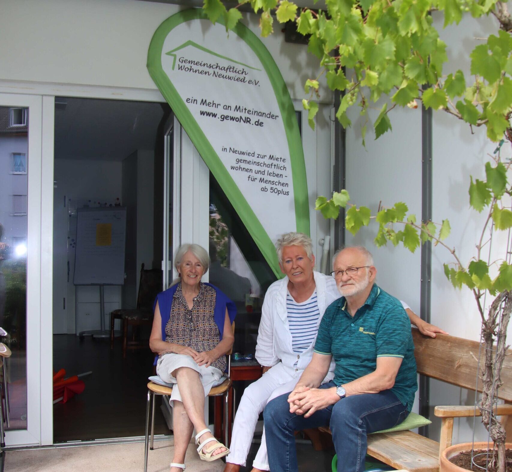 Drei Personen sitzen entspannt vor einer Infofahne zum gemeinschaftlichen Wohnen und Leben in einer Mieterwohnung
