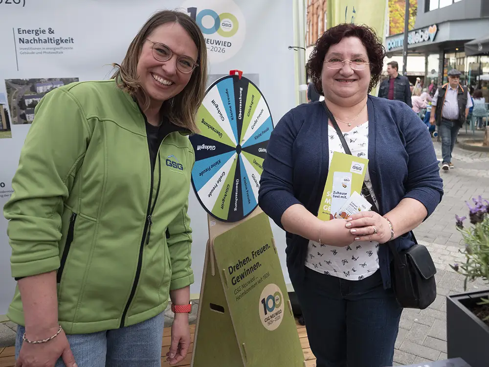 R_Claassen-1044636 Zwei freundlich lächelnde Frauen stehen vor einem GSG Pavillon mit Glücksrad und Informationsmaterial zum Mitnehmen