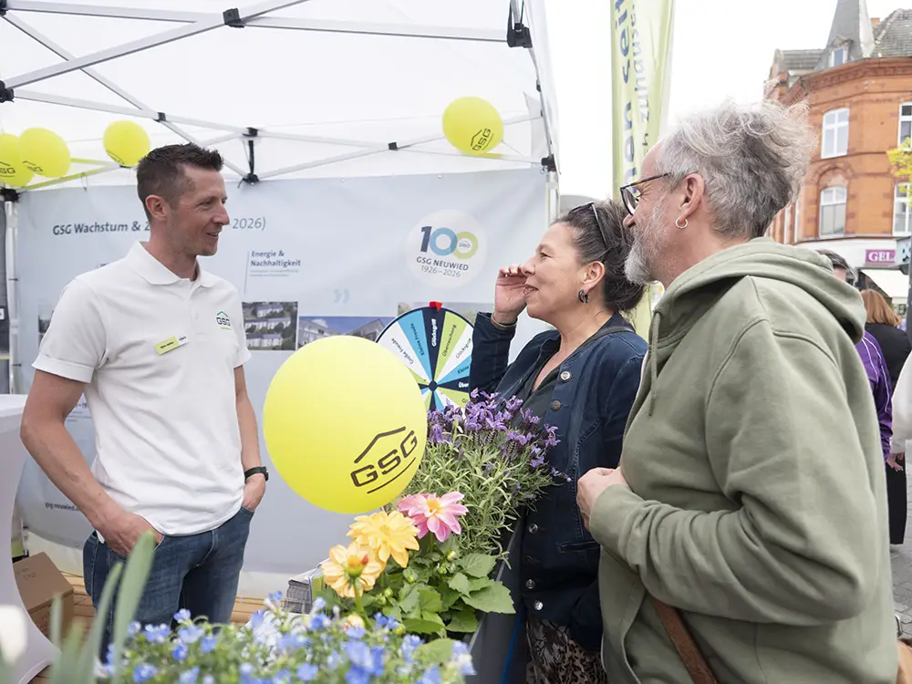 Ein GSG-Mitarbeiter im Gespräch mit Besuchern an einem Informationsstand mit gelben Luftballons und einem Glücksrad