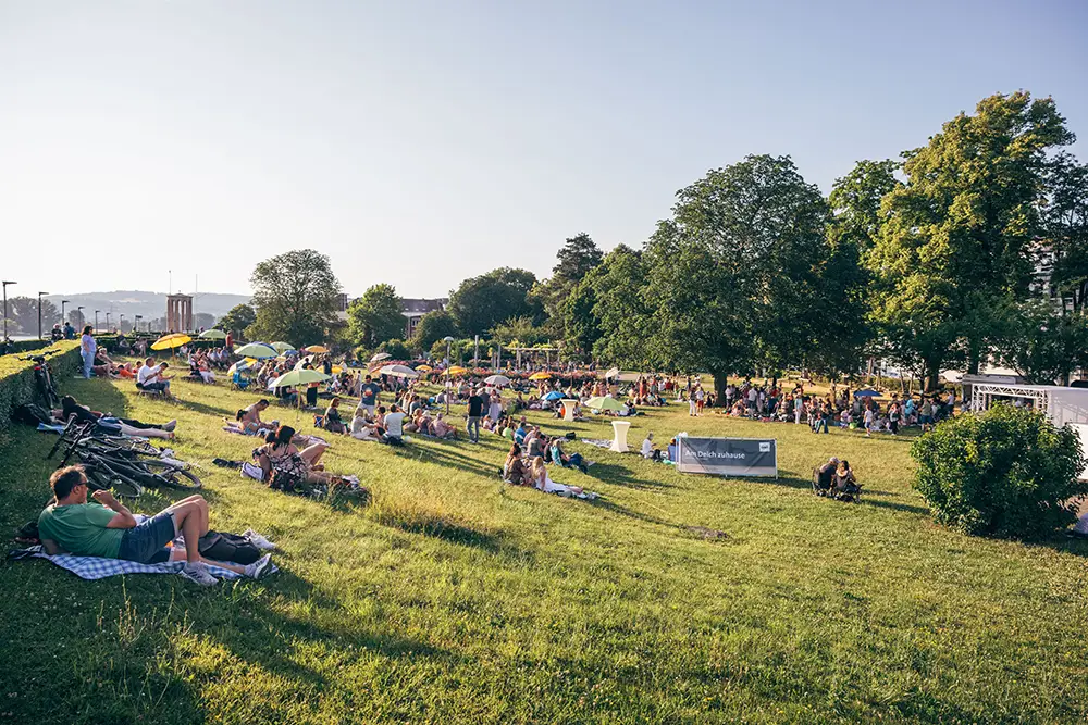 Viele Menschen entspannen beim Picknick auf einer sonnigen Wiese am Deich mit bunten Sonnenschirmen