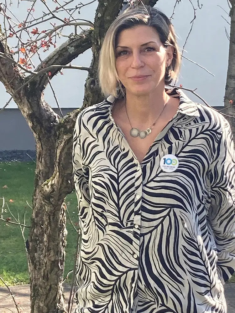 Alexandra_Gras Alexandra Gras mit blondierten Haaren in einer schwarz-weiß gemusterten Bluse vor einem Baum stehend