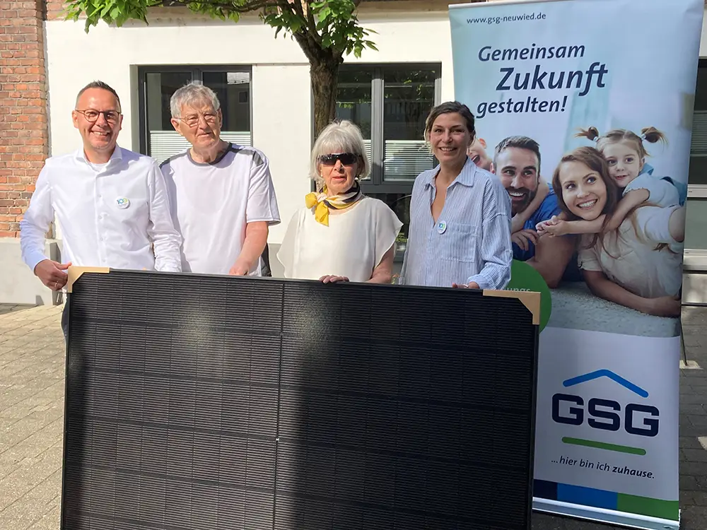 Vier Personen halten gemeinsam ein schwarzes Balkonkraftwerk bei einer feierlichen Übergabe vor einem GSG Banner