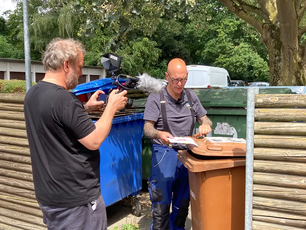 Ein Kameramann filmt Franz Humer bei der Kontrolle von Mülltonnen im Freien