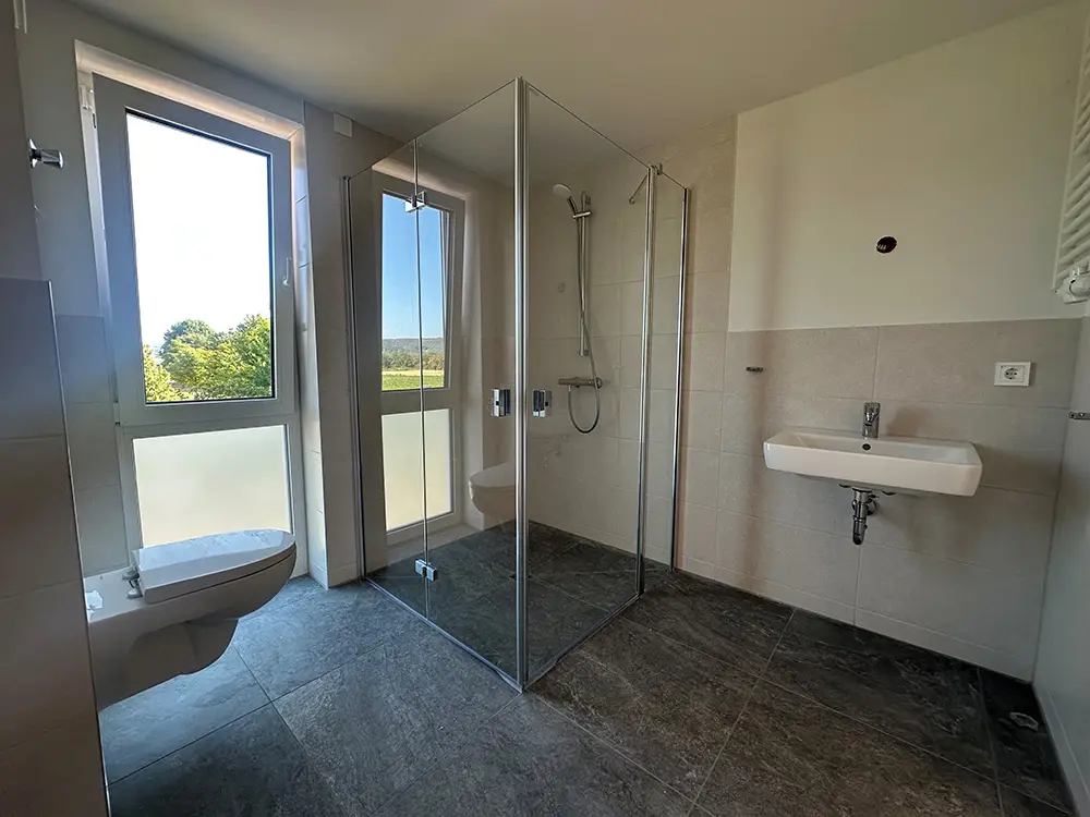 Bad Helles Badezimmer einer Neubauwohnung mit bodengleicher Glasdusche, Wandwaschbecken und Fensterblick ins Grüne