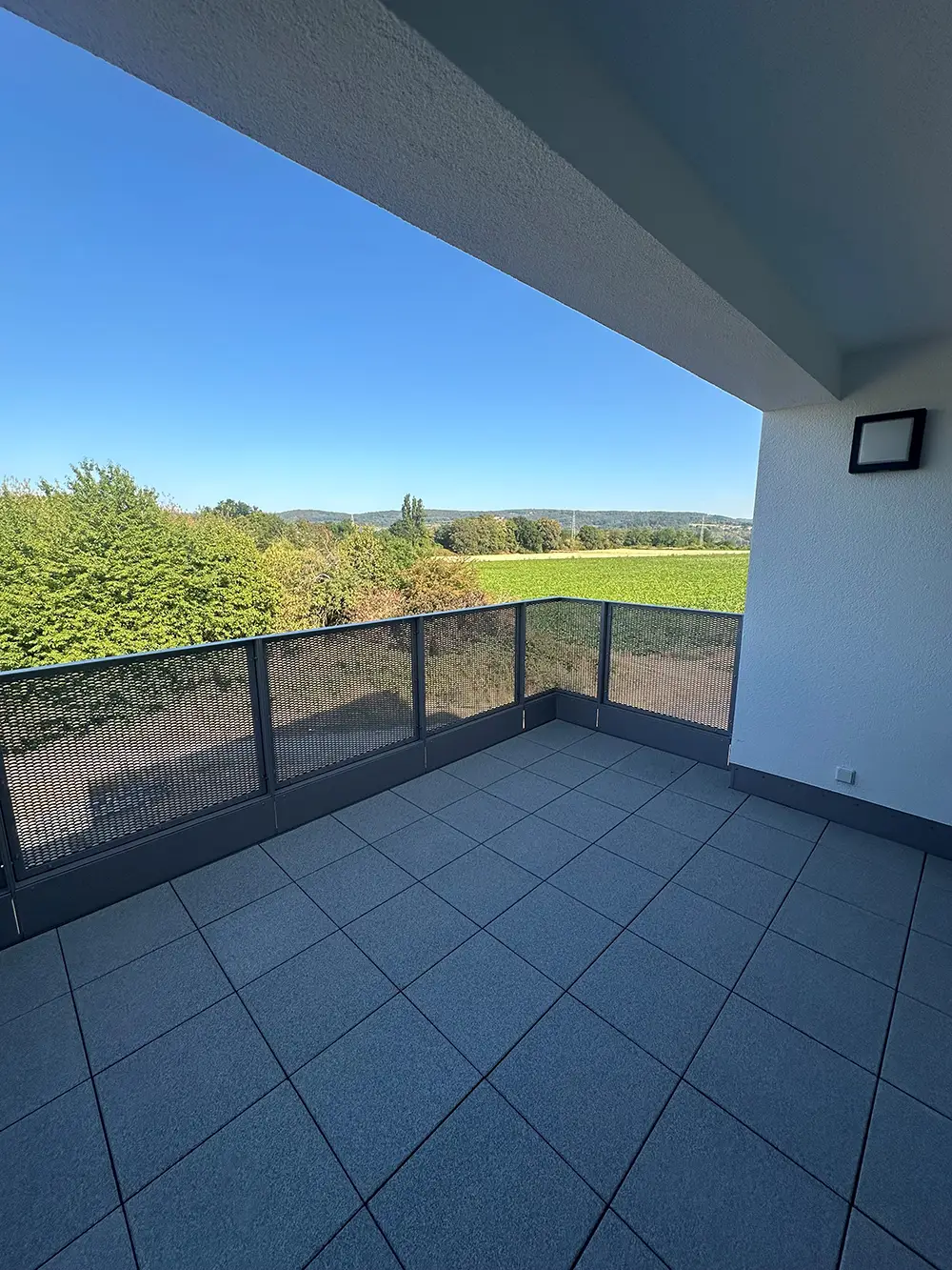 Balkon Weitläufiger Balkon einer Neubauwohnung mit grauen Fliesen, Metallgeländer und freiem Blick in eine grüne Landschaft