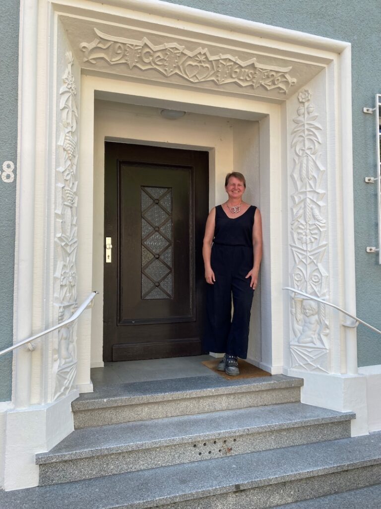 GSG saniert gechütztes Haus d Eine Frau steht lächelnd vor der sanierten Eingangstür eines geschützten Hauses mit kunstvollem Steindekor
