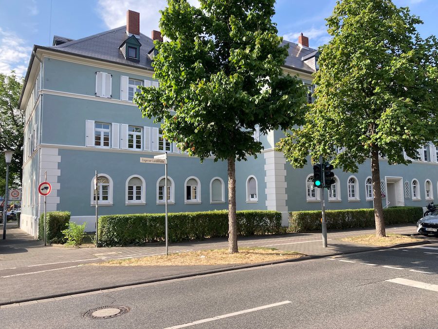 Ein hellblau saniertes denkmalgeschütztes Wohngebäude mit weißen Fensterläden hinter grünen Bäumen an einer Straße