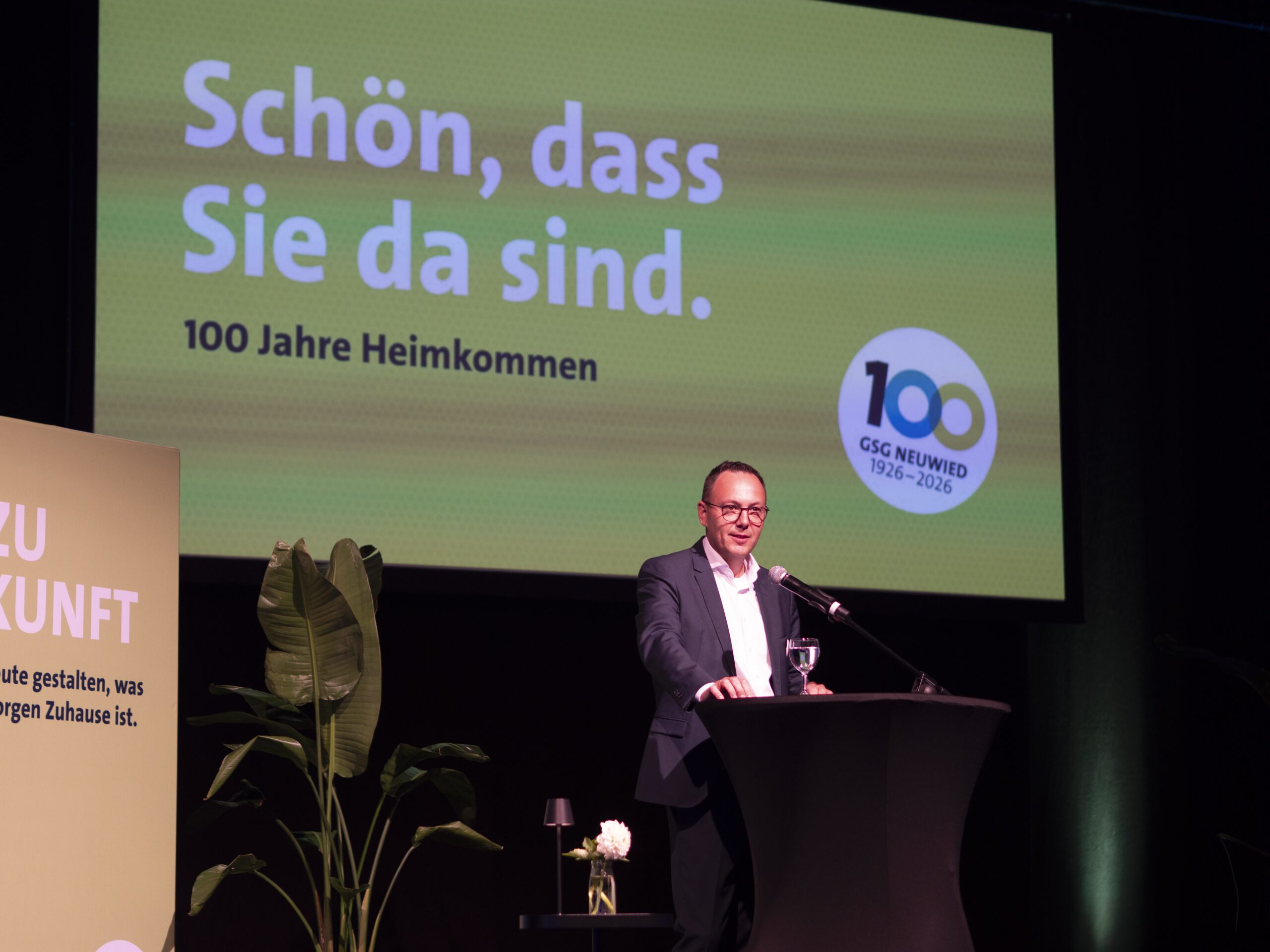 Ein Mann spricht bei einer Jubiläumsveranstaltung zu 100 Jahren vor einer Leinwand mit der Aufschrift 100 Jahre Heimkommen