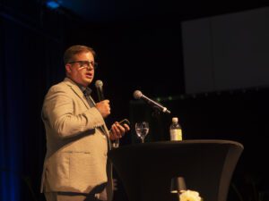 R_Claassen-1055423 Ein Brillenträger im hellen Anzug hält bei einer Jubiläumsveranstaltung mit Mikrofon in der Hand einen Vortrag