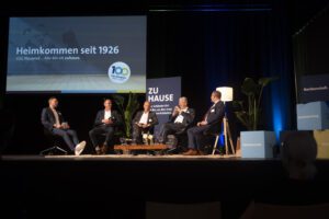 R_Claassen-1788327 Fünf Männer sitzen auf einer Bühne in Sesseln für eine Podiumsdiskussion vor einer Leinwand mit Jubiläumslogo
