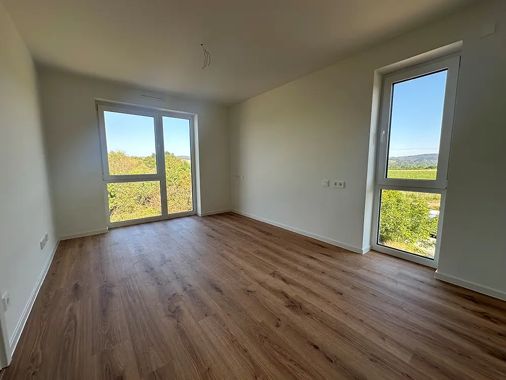Wohnung-rechts-hinten Leerer Wohnraum einer Neubauwohnung mit hellem Laminatboden und zwei großen Fenstern mit Ausblick ins Grüne