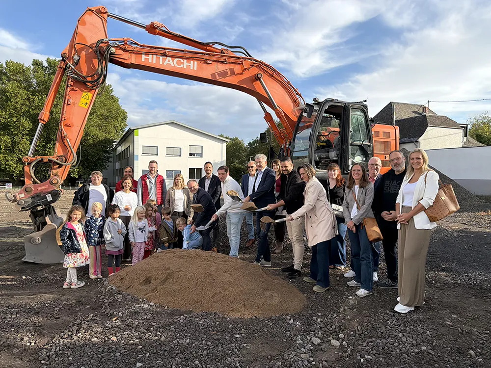 Erster Spatenstich: Gruppe mit Kindern und Bagger bewegt Erde auf gravel Fläche