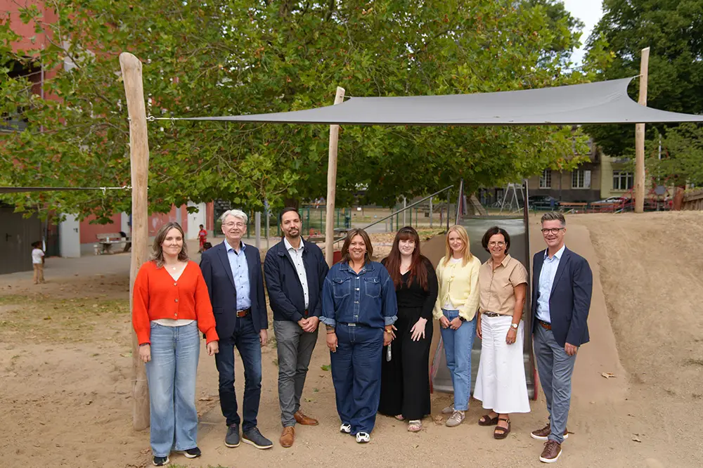 Eine Gruppe von Erwachsenen steht unter einem neuen Sonnensegel auf einem Spielplatz mit Kinderschiff