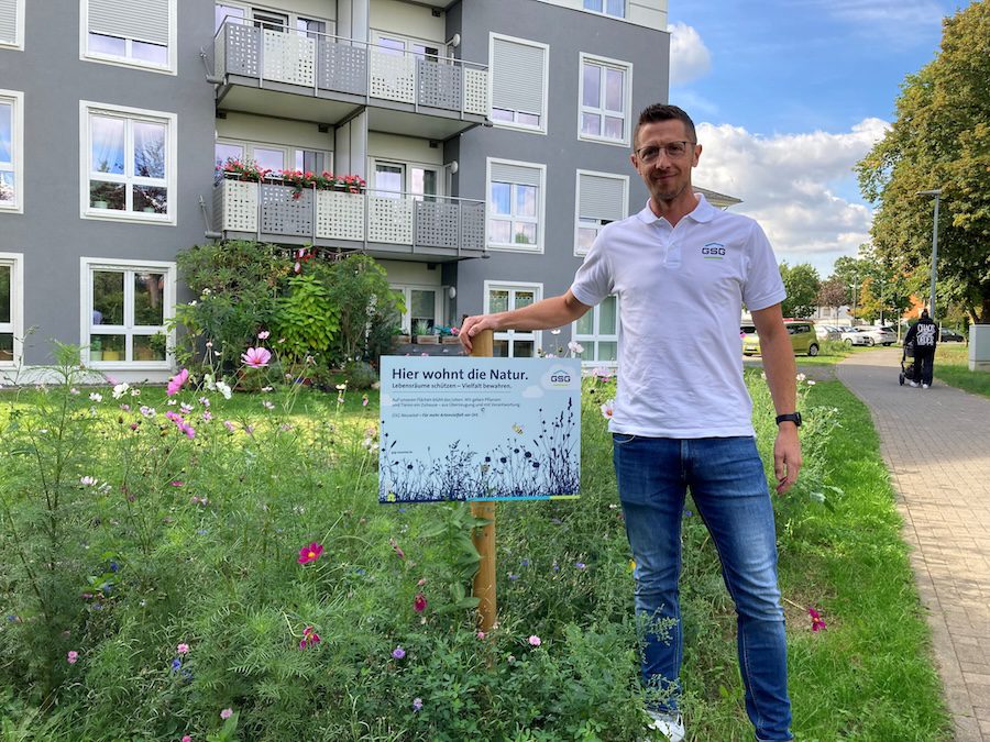 GSG-Mitarbeiter Björn Müller steht neben einem Hinweisschild 'Hier wohnt die Natur' vor einer Biodiversitätsfläche mit Blumen und Gräsern.